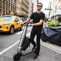 Man holding a scooter on a city street with a taxi in the background
