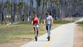 Two people riding electric scooters on a path with palm trees in the background