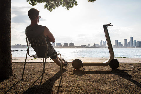 Person sitting on a chair with a scooter next to them, overlooking a city skyline.