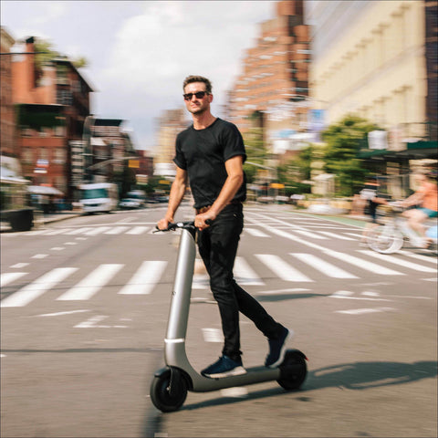 Man riding a electric scooter on a city street with buildings and pedestrians in the background