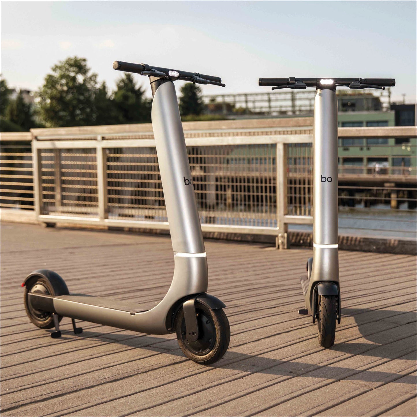 Two electric scooters on a wooden deck with a bridge and trees in the background.
