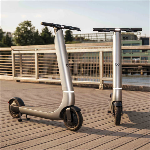Two electric scooters on a wooden deck with a bridge and trees in the background.