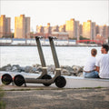 Two people sitting on a bench by a waterfront with an electric scooter nearby, city skyline in the background.