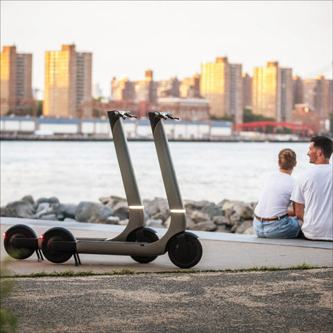 Two people sitting on a bench by a waterfront with an electric scooter nearby, city skyline in the background.