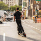 Person riding an electric scooter on a city street with buildings and traffic in the background