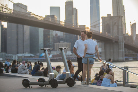 Two electric scooters with gray frames and black wheels are parked on a waterfront promenade in New York City, with the Brooklyn Bridge and city skyline in the background. Two people are standing nearby, and other people are seated along the railing.